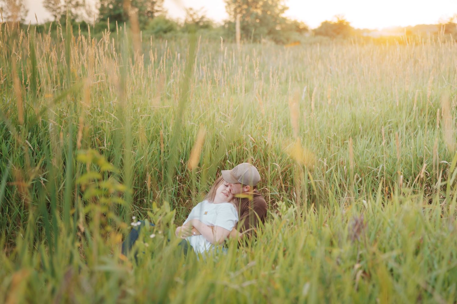 Couple sitting closely together in tall golden grass during a warm sunset.