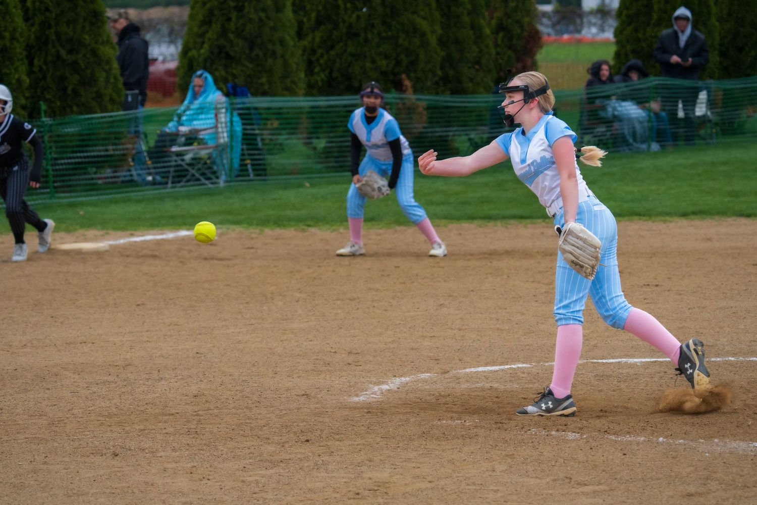 Female softball pitcher in light blue uniform delivers a pitch during an overcast outdoor game with a teammate in the background.