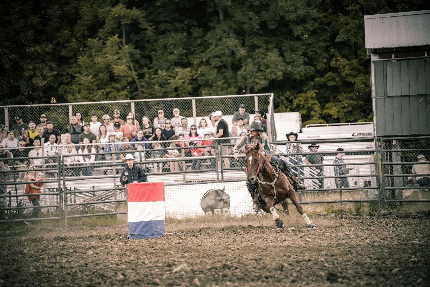Cowboy on horseback racing past a Dutch flag in a dirt rodeo arena surrounded by spectators and trees.