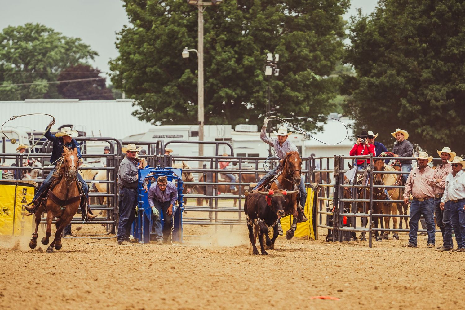 Cowboys compete in a lively rodeo event with riders on horseback and a bull in a sandy arena surrounded by spectators.
