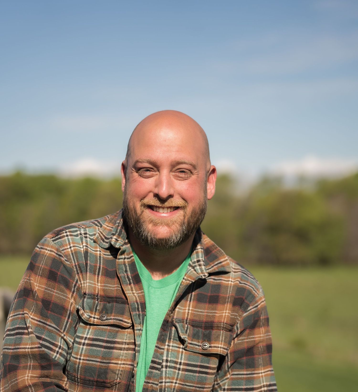 Smiling bald man with beard wearing plaid shirt outdoors in green field under blue sky.