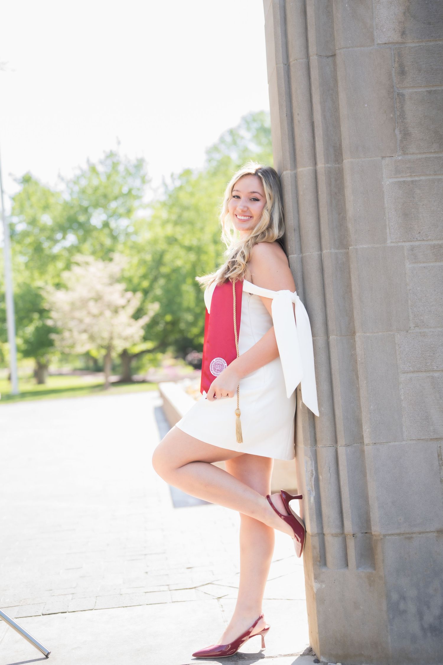 Smiling blonde woman in white dress holding red graduation regalia, leaning against stone column outdoors.