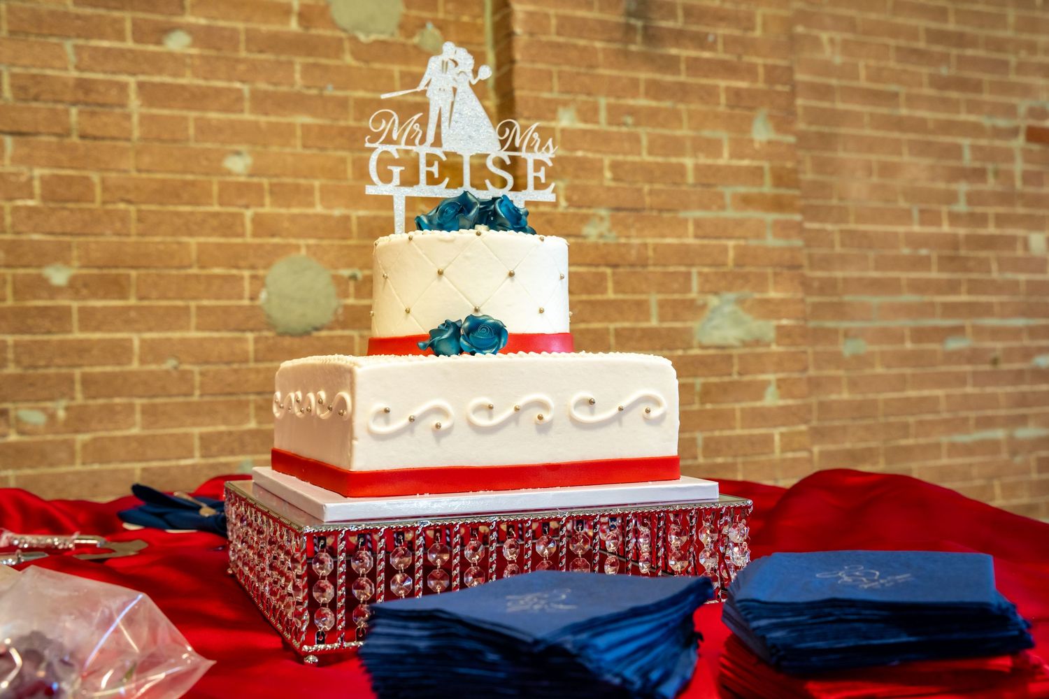 Elegant three-tier wedding cake with teal flowers and Mr. & Mrs. Geise topper on red table.