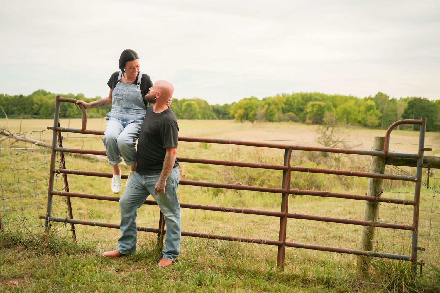 Man helping woman climb a rustic farm gate in an open countryside field.