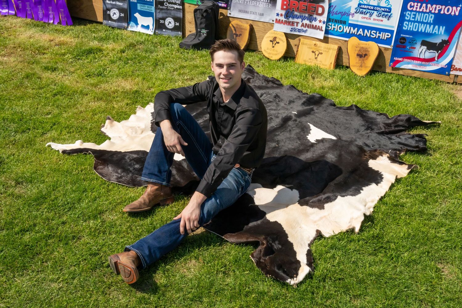 Young man sitting casually on a black and white cowhide rug outdoors with show banners behind him.