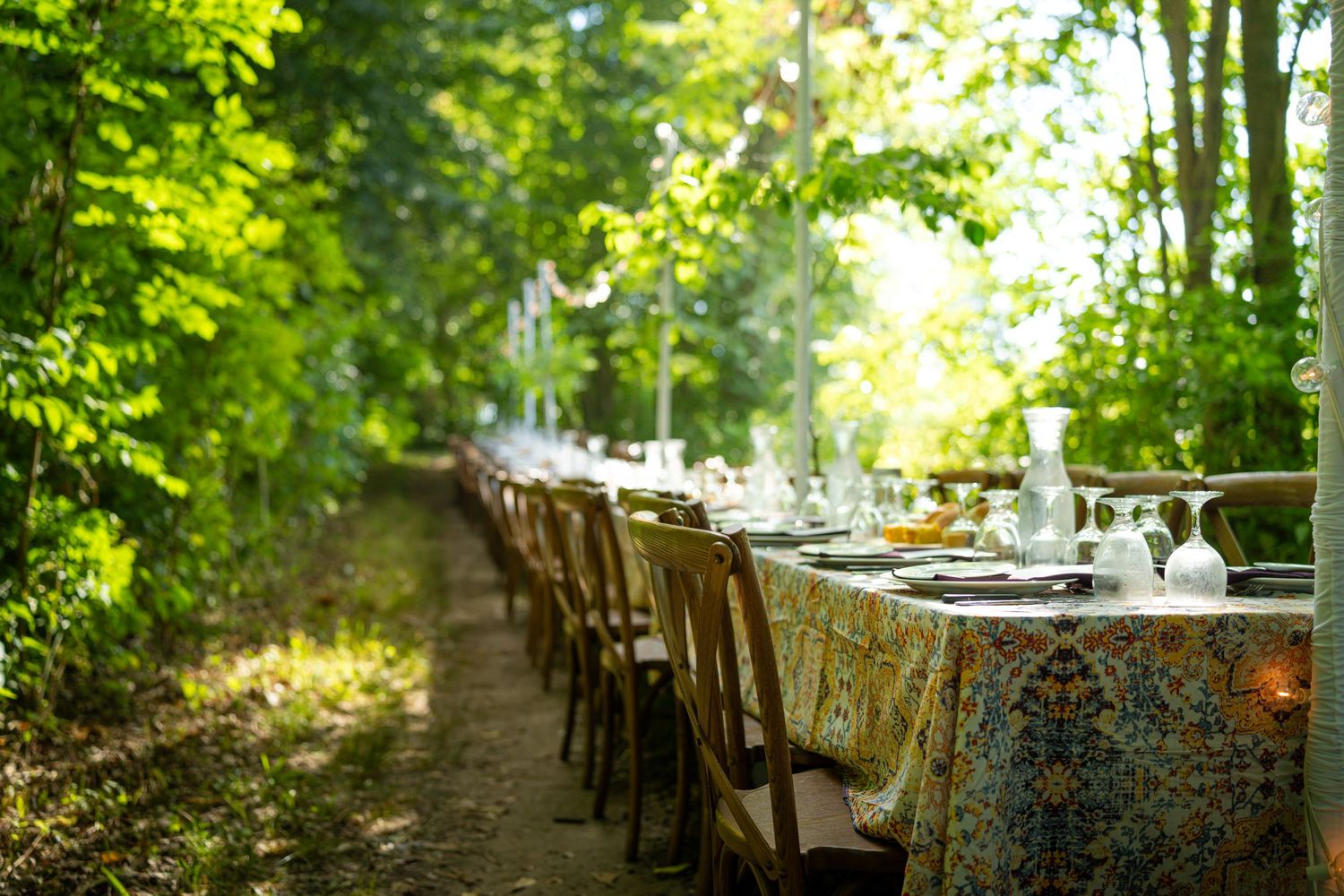 Long outdoor banquet table with floral tablecloth set for dining along a tree-lined garden path.