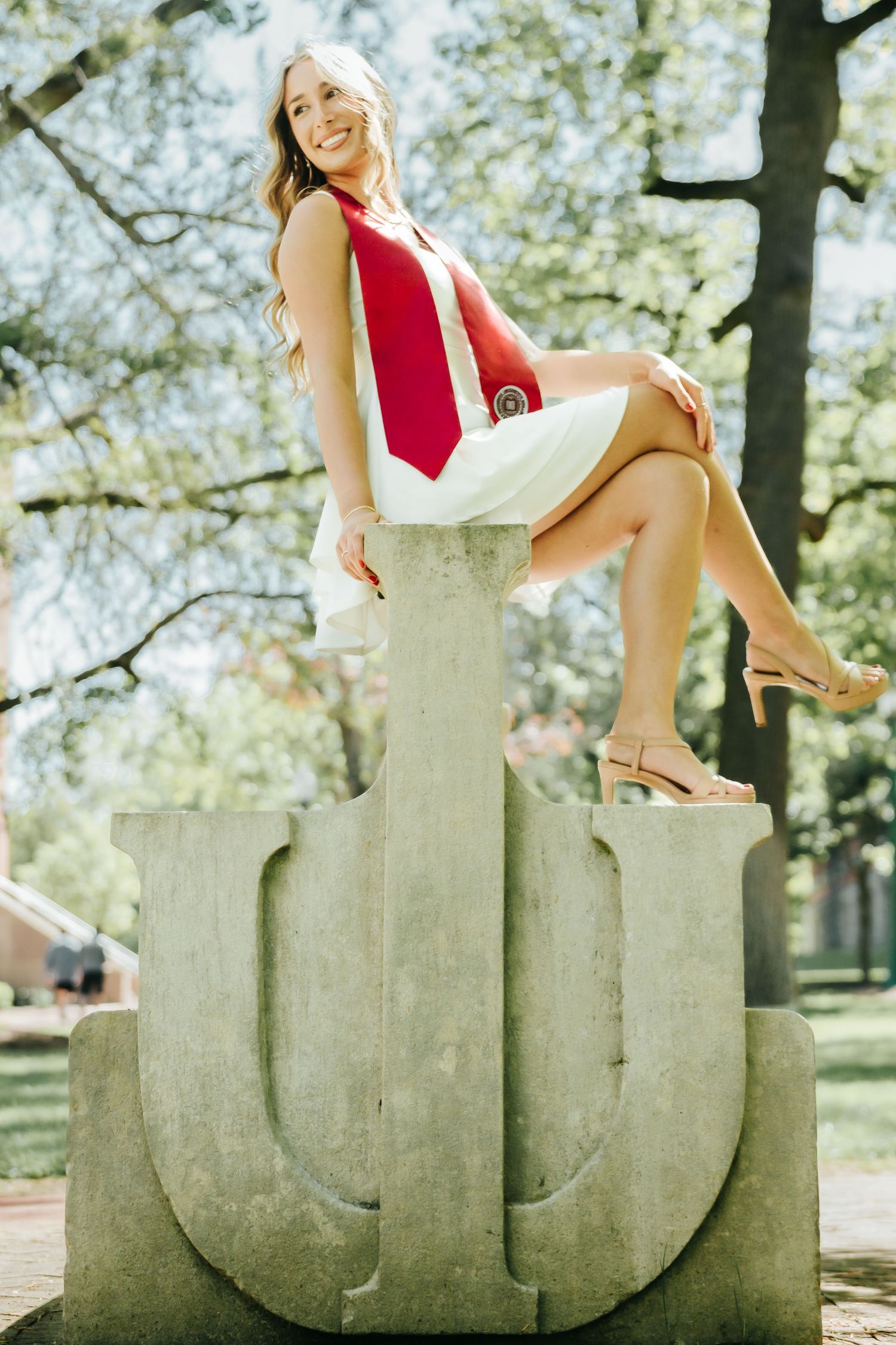 Smiling blonde graduate in white dress sitting on Indiana University stone IU sculpture outdoors.