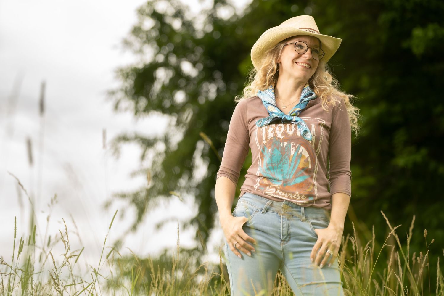 Young woman in cowboy hat, glasses, and jeans smiling outdoors in a grassy field with trees in background.