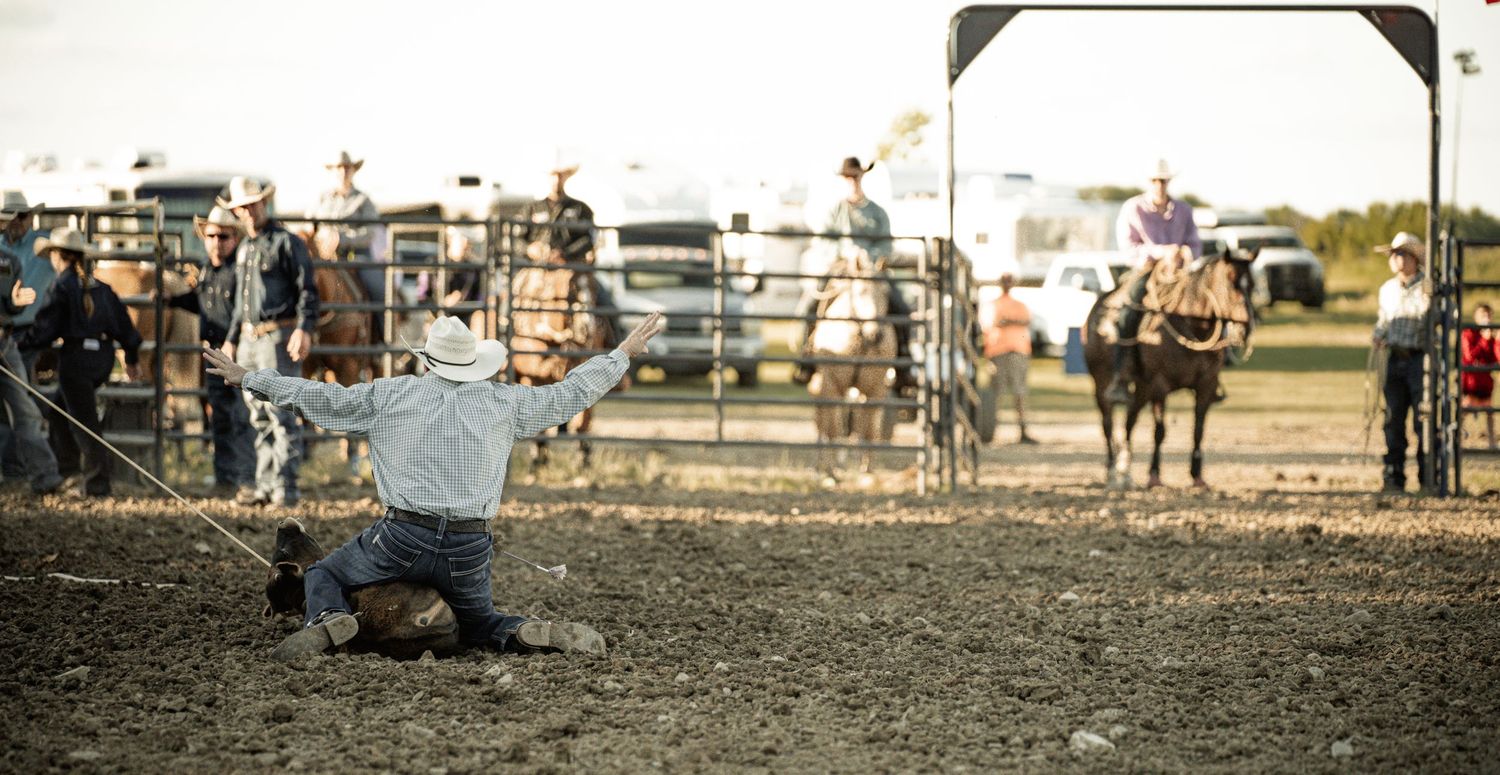 A cowboy kneels in the dirt after roping an animal during a competitive rodeo tie-down roping event.