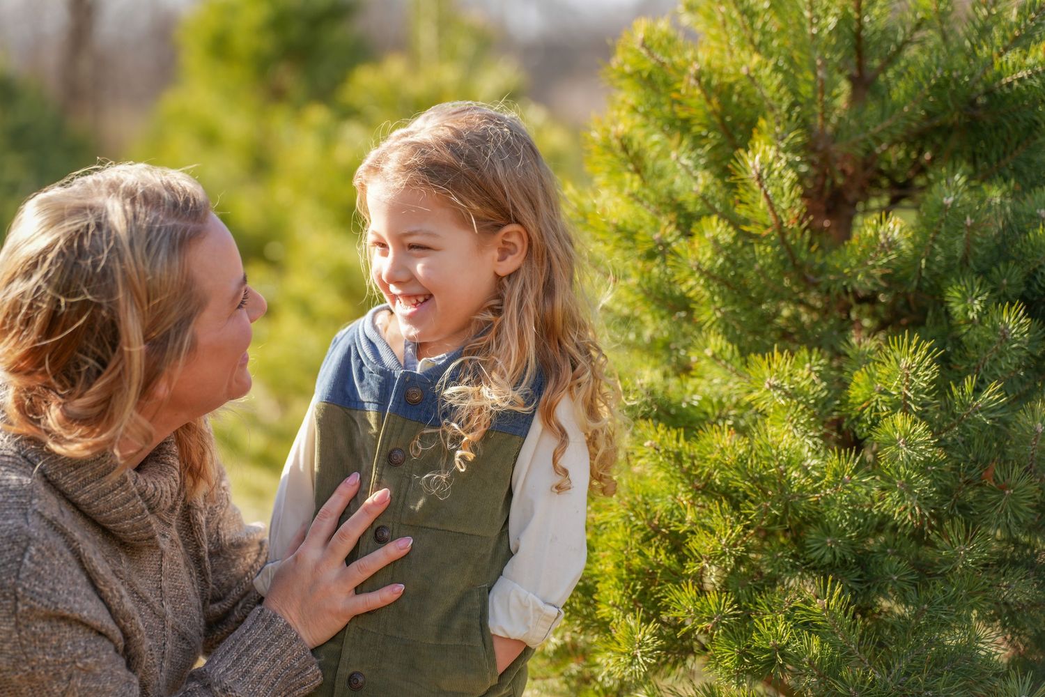 A woman crouches beside a smiling young girl near a pine tree at a Christmas tree farm.