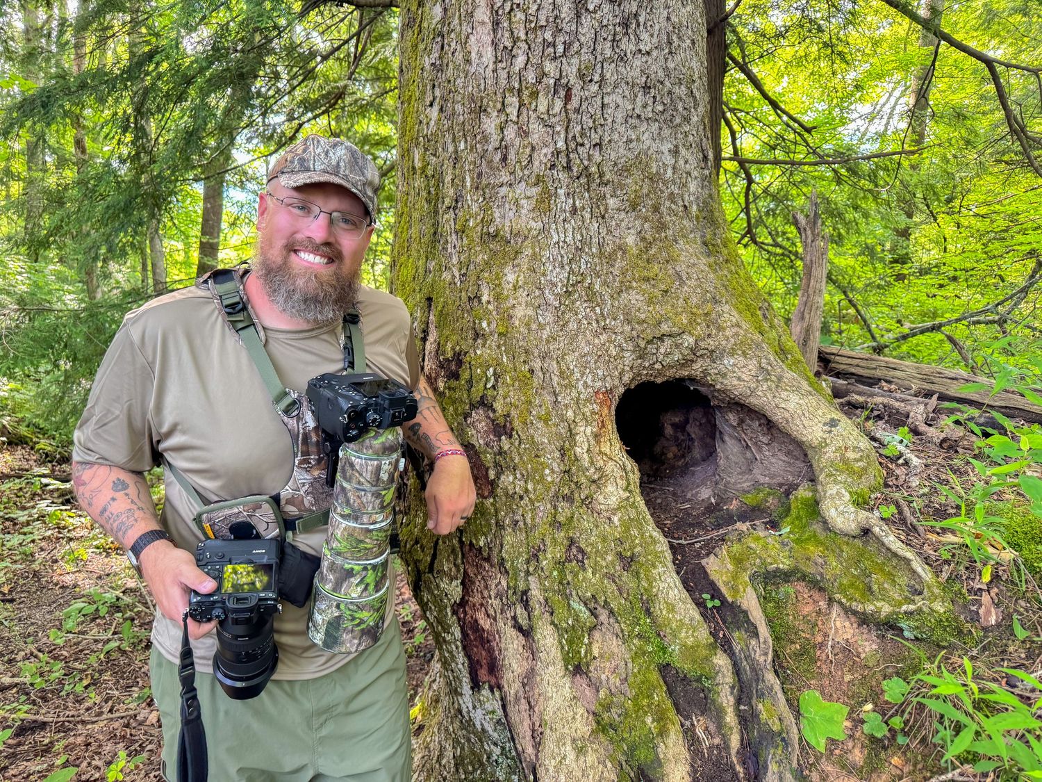 Bearded man with cameras and camo lens stands beside large tree with hollow in lush green forest.