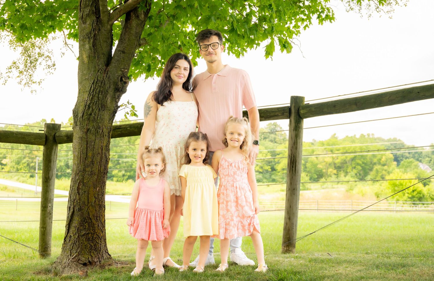 Family of five posing outdoors near a large tree and wooden fence on a lush green lawn.