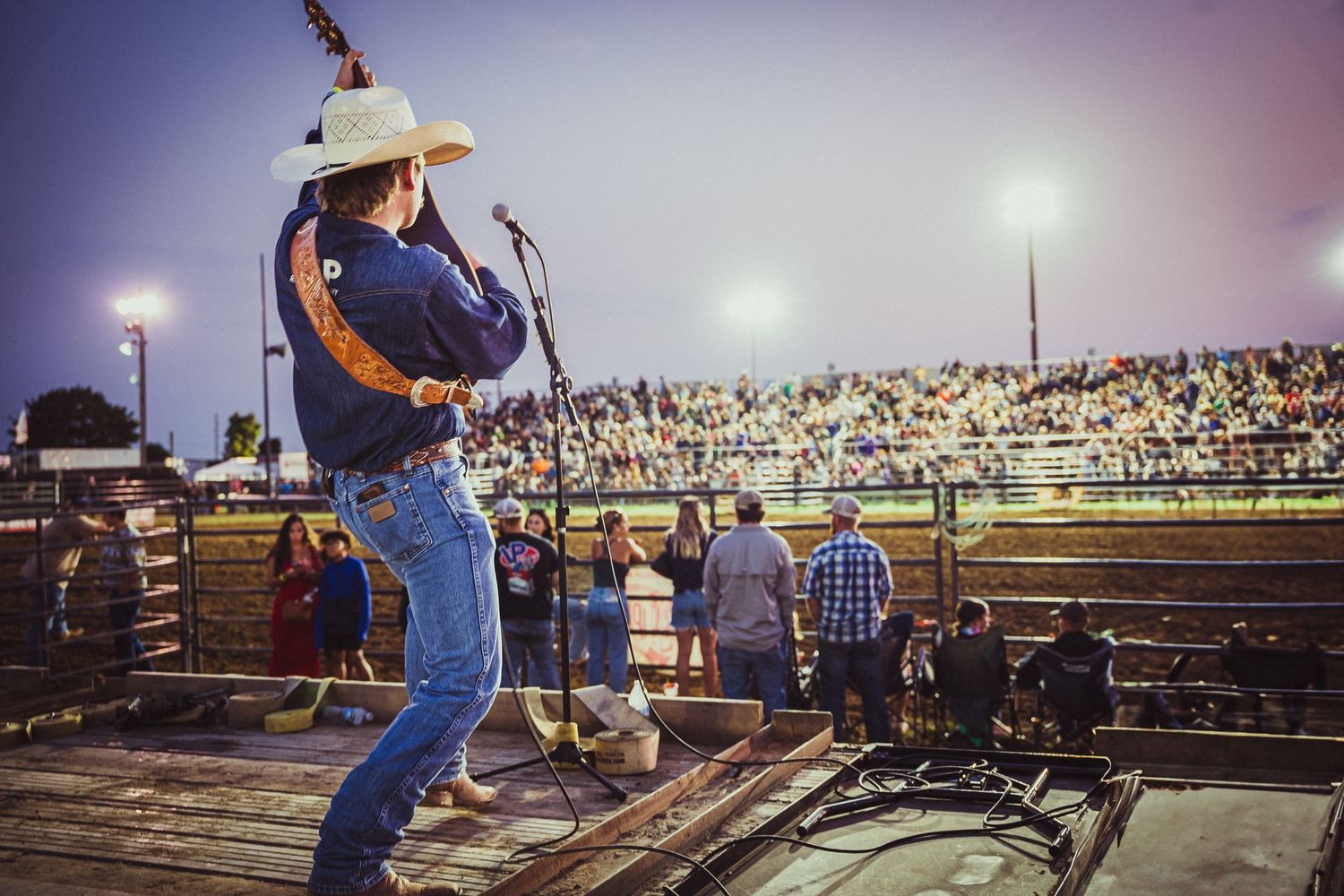 Country singer performs on stage at a rodeo arena packed with fans under evening sky.