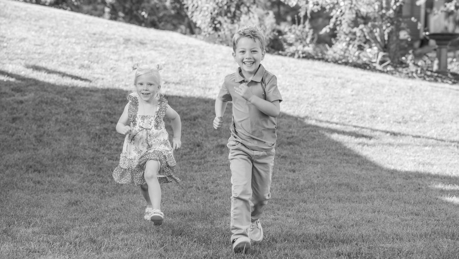 Two young children laughing and running together on green grass in a black and white outdoor photo.