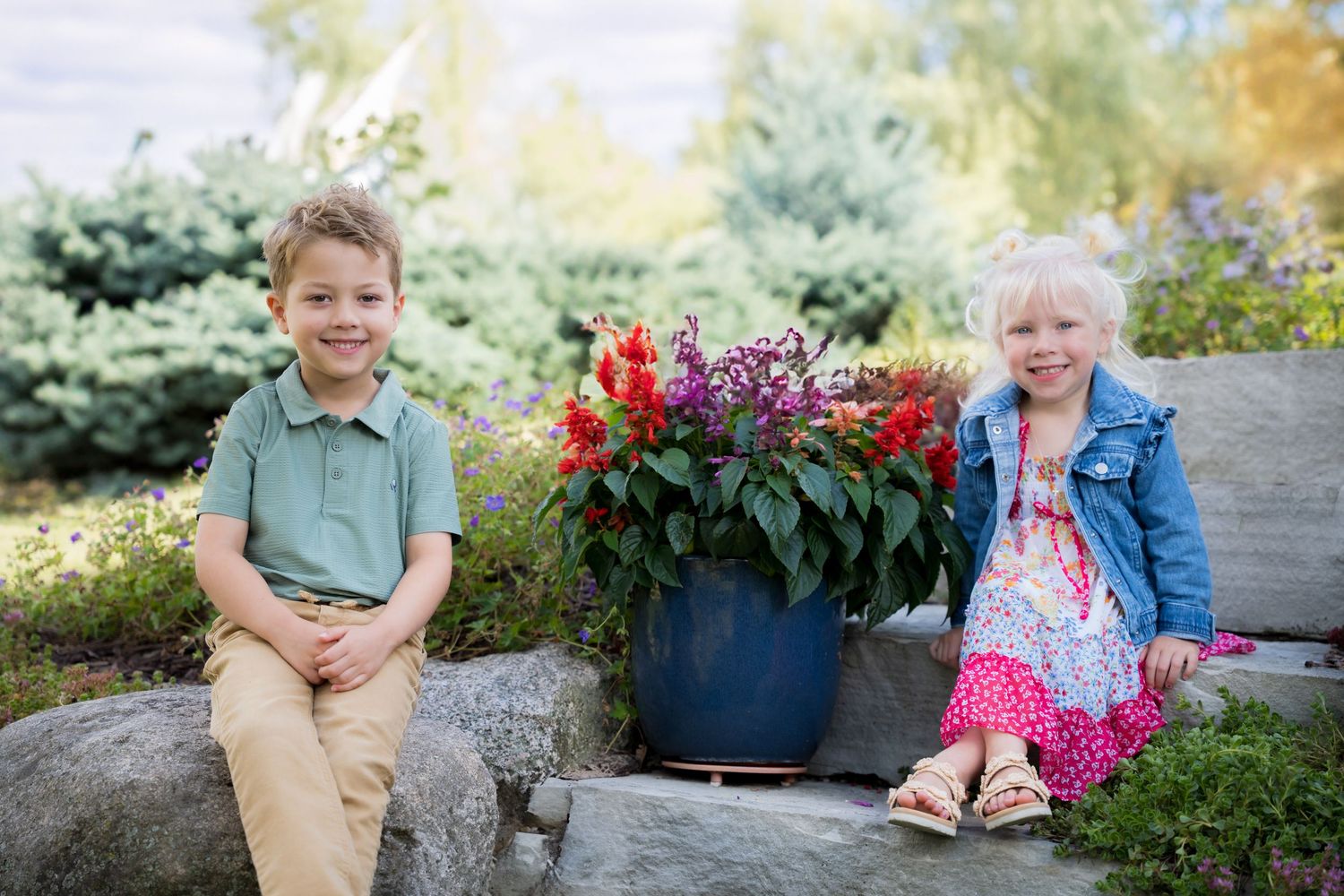 Two young children sitting on rocks beside a colorful flower pot in a lush garden setting.