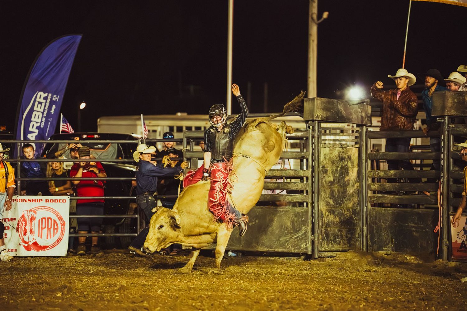 A bull rider competes at a nighttime rodeo event, with the bull bucking dramatically in a dirt arena.