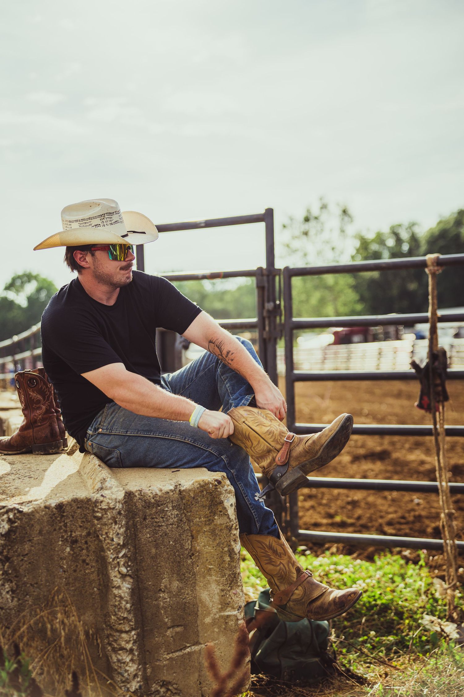 A cowboy in a white hat and jeans sits on a rock near a ranch fence, wearing boots and a black t-shirt.