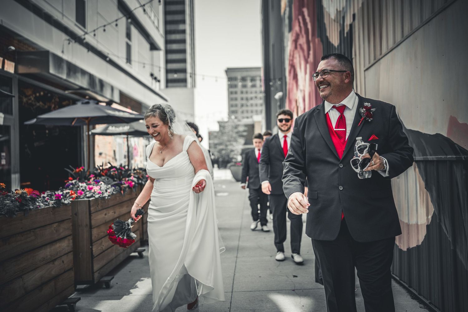 Bride and groom walking down urban sidewalk with groomsmen in red ties, city buildings in background.