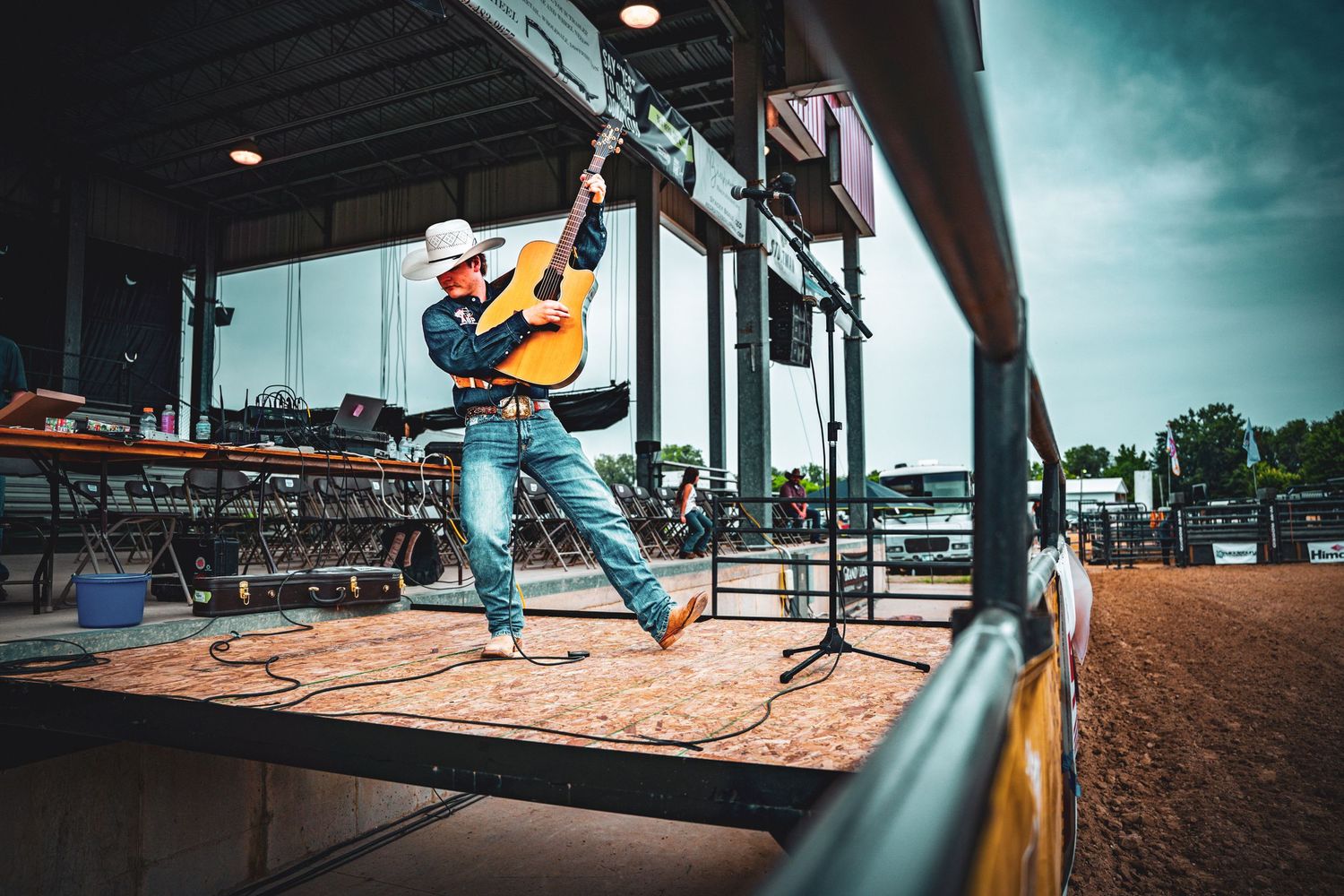 Country musician in cowboy hat plays guitar on outdoor stage at a rodeo arena venue.
