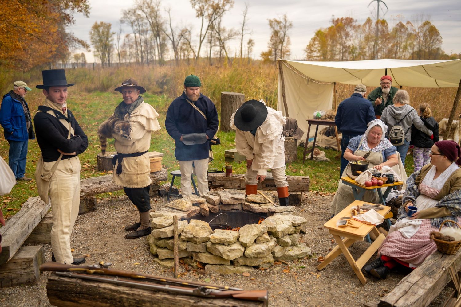 Historical reenactors cook over a stone fire pit in period costumes during an outdoor autumn encampment.