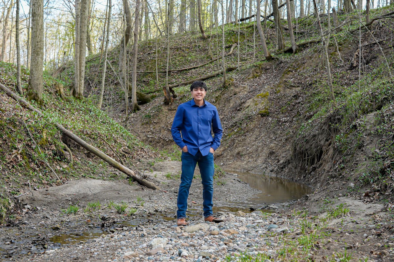 Young man in blue jacket standing in a wooded ravine with a small stream on a spring day.