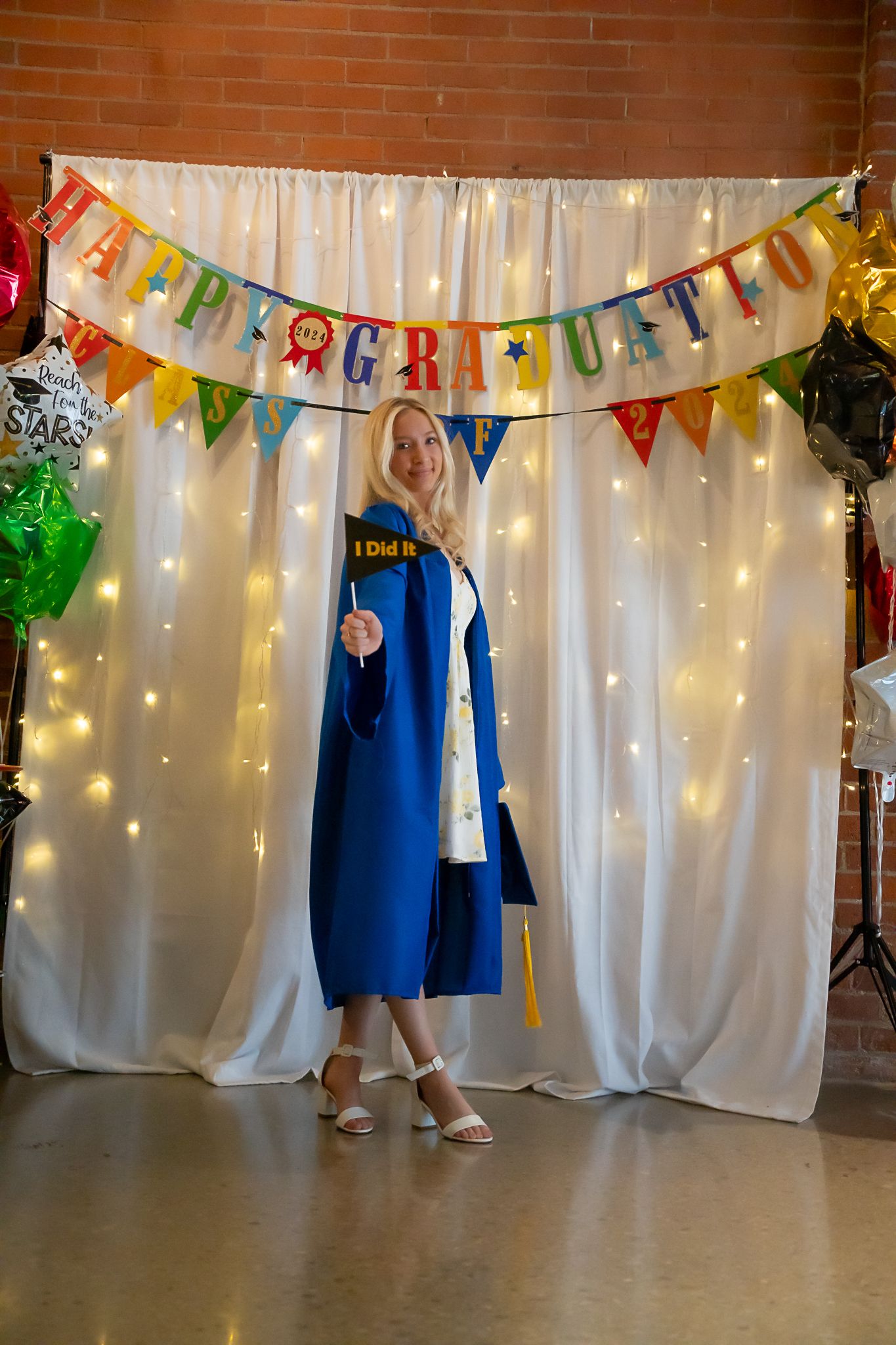 Blonde graduate in blue gown posing before Happy Graduation banner with fairy lights and balloons.