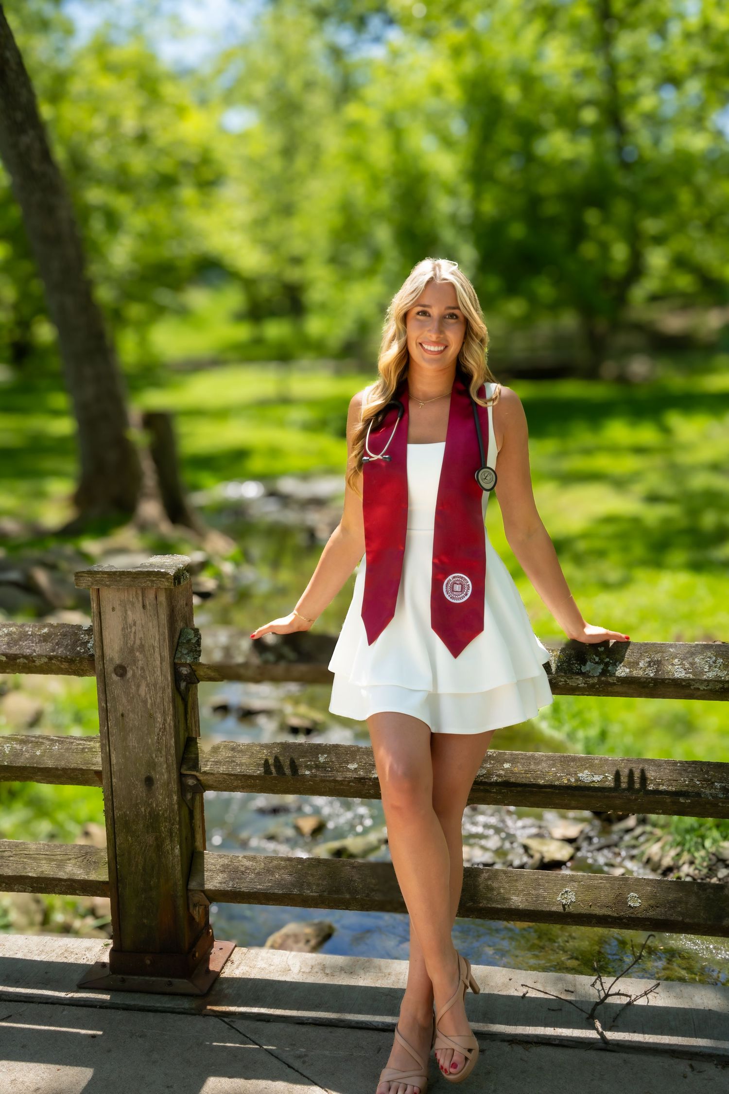 Smiling blonde nursing graduate with stethoscope and red stole posing on wooden bridge outdoors.