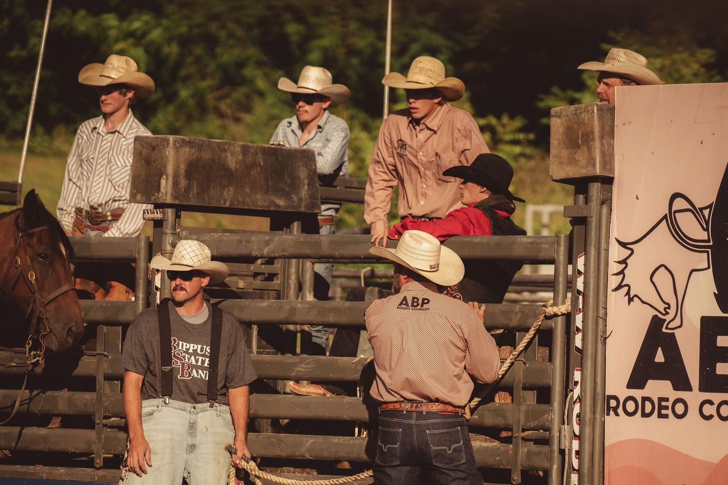 Group of cowboys in white hats gather around rodeo chutes near an AEP Rodeo Company sign at dusk.