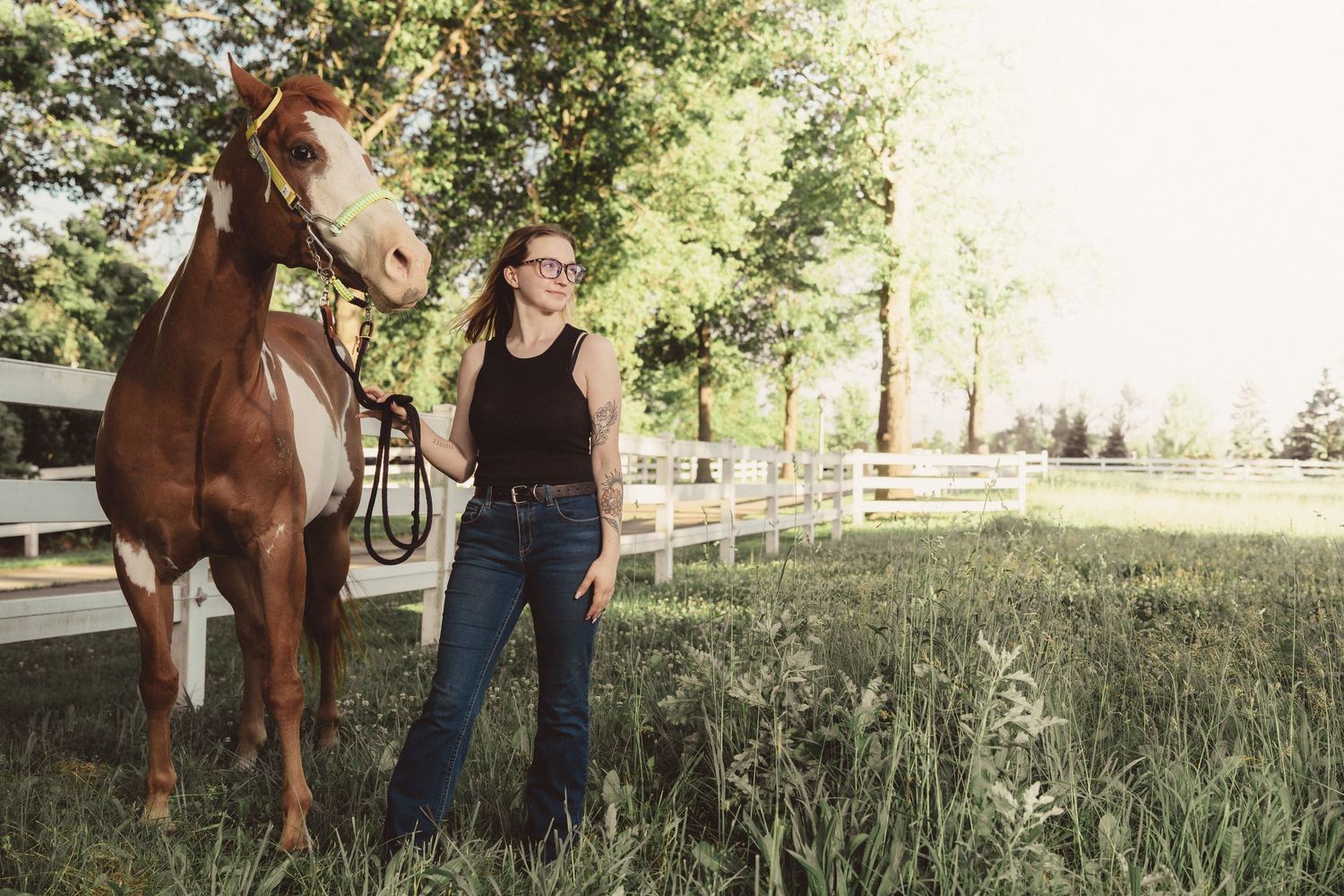 Woman in black top and jeans stands beside a chestnut horse in a lush green pasture with white fence.