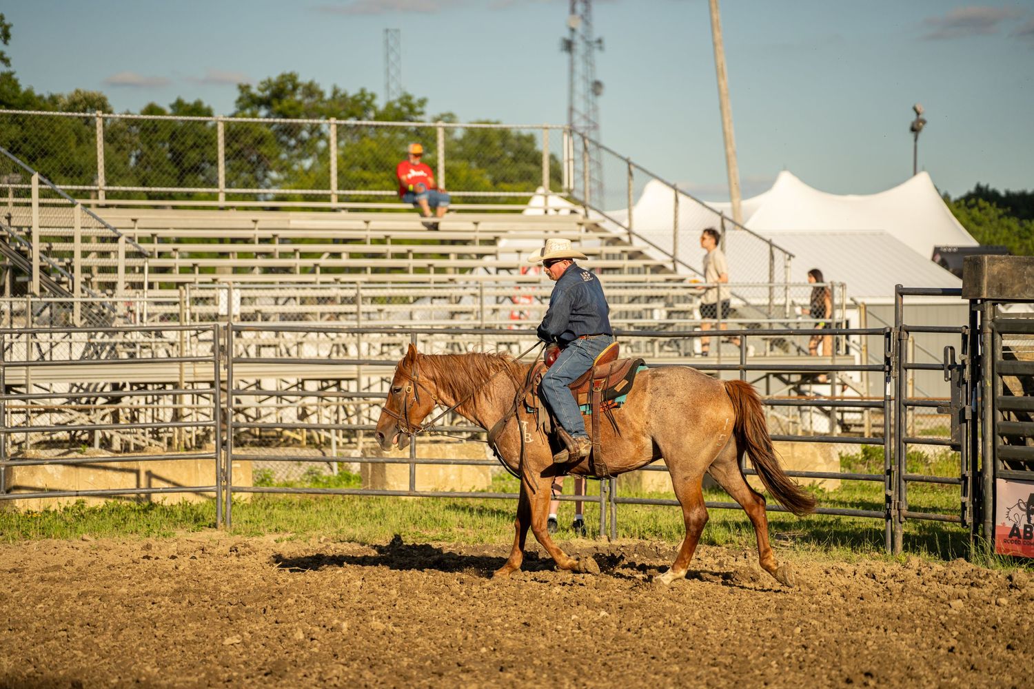 A cowboy rides a tan horse in an arena with bleachers and a white tent in the background.