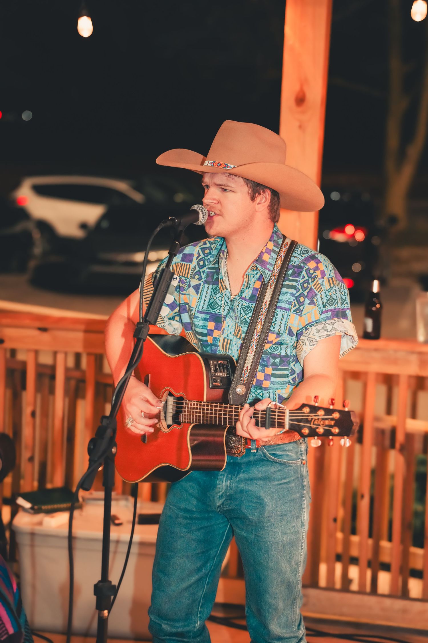 Young country singer in cowboy hat and tropical shirt performs with red acoustic guitar at outdoor night venue.