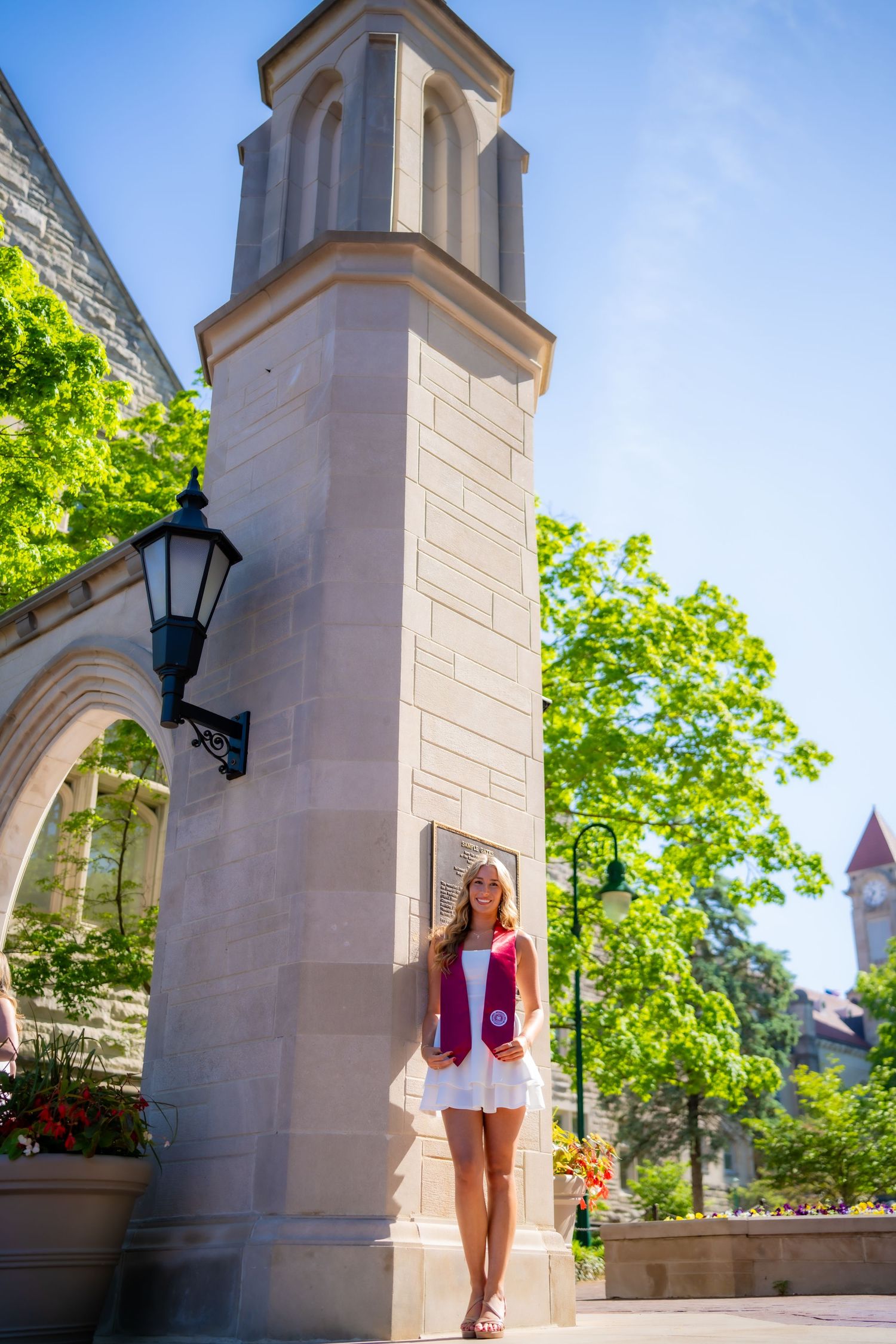 Female graduate in white dress with IU crimson stole stands beside ornate stone campus building.