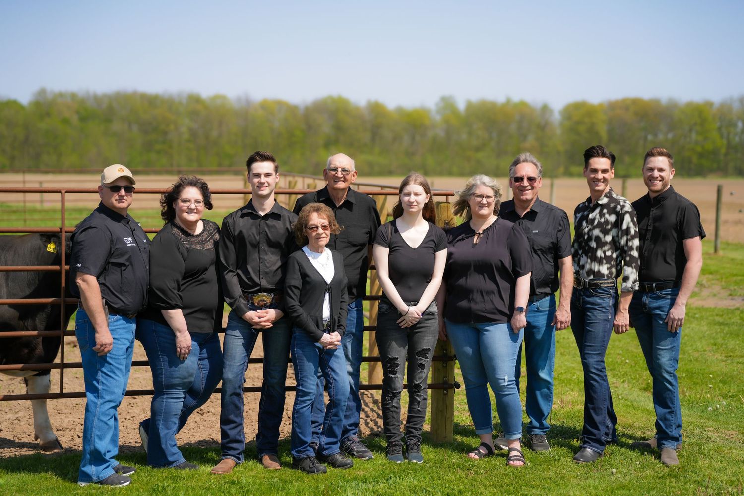 Group of ten people smiling together outdoors near a farm gate on a sunny spring day.