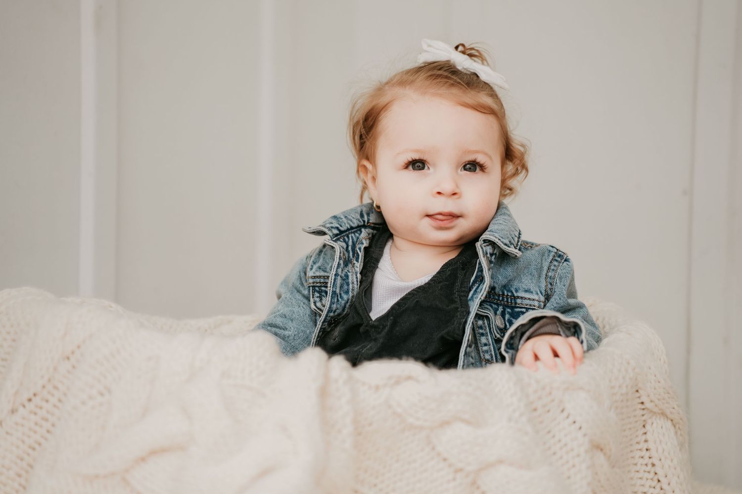 An adorable baby girl in a denim jacket sits on a knit blanket looking at the camera.
