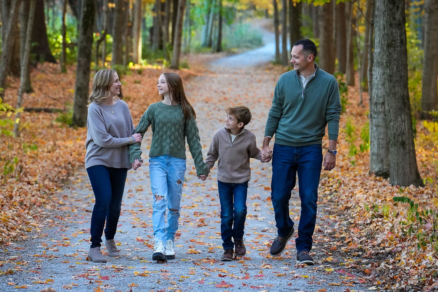 A family of four walks hand in hand down a leaf-covered path through a golden autumn forest.