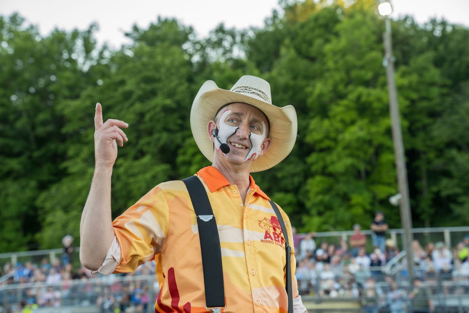 A rodeo clown in an orange shirt with face paint points upward while entertaining the crowd at a rodeo.