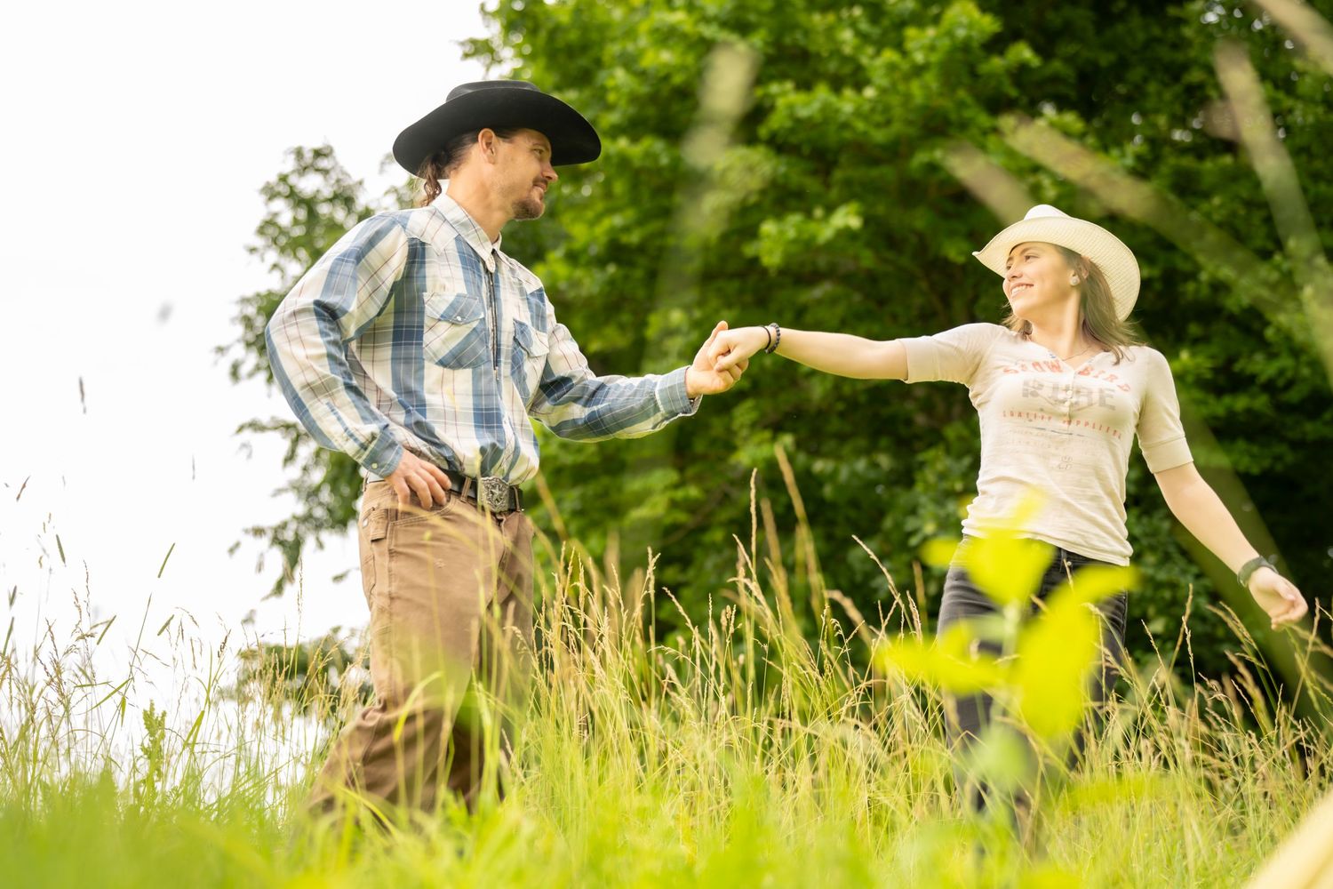A couple in cowboy hats hold hands and dance in a lush green meadow on a sunny day.