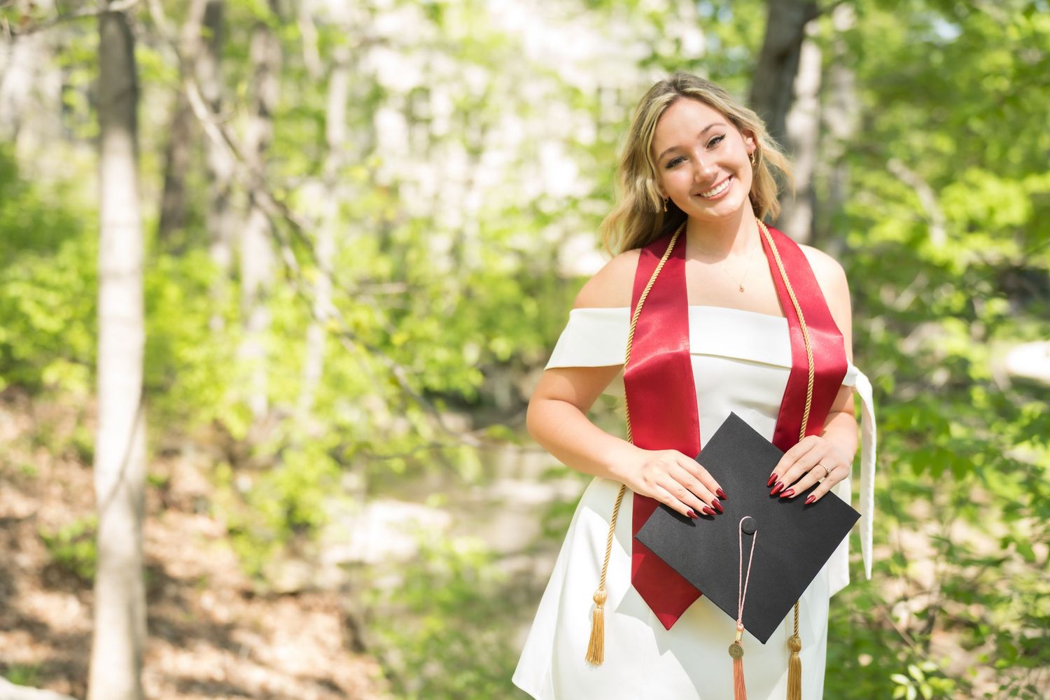 Smiling female graduate in white dress with red sash holds cap outdoors among green trees on sunny day.