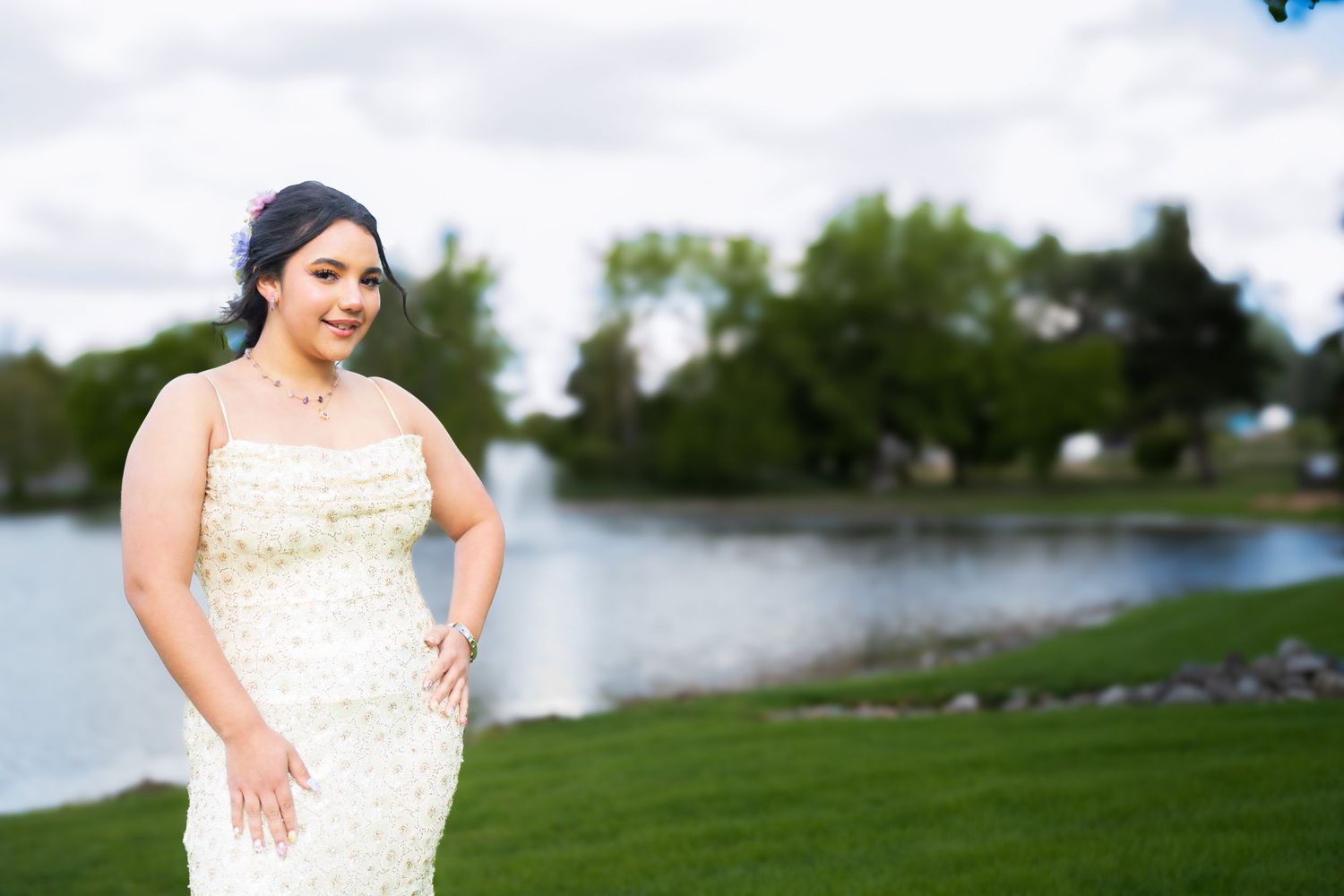 Woman in beaded white gown posing outdoors near a serene pond with lush green grass and trees in the background.