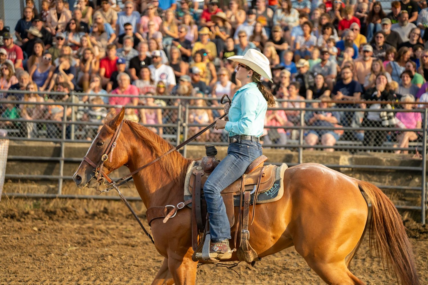 A cowgirl in a white hat rides a chestnut horse at a rodeo arena packed with spectators.