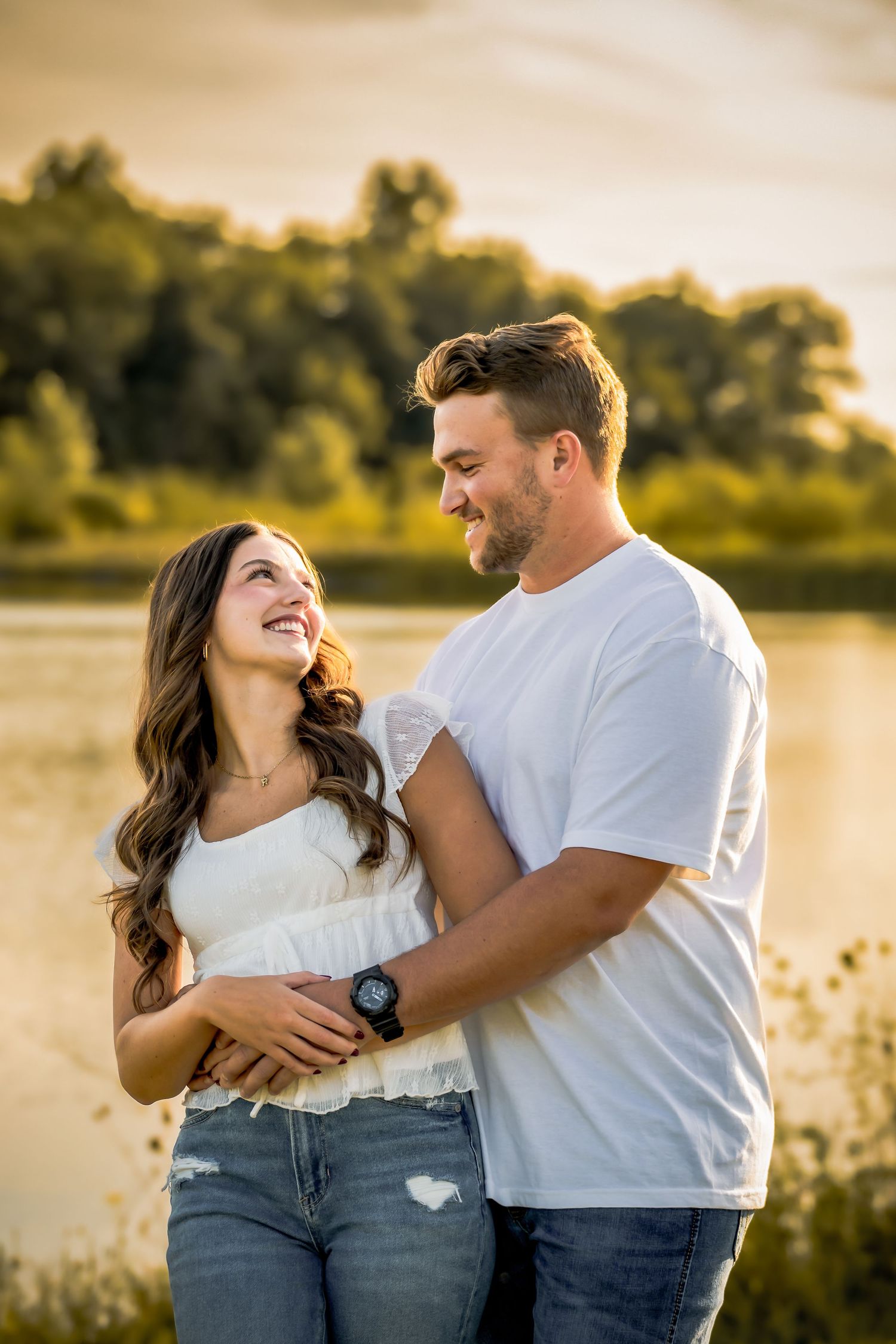 A happy young couple embraces and smiles at each other near a golden sunset lake backdrop.