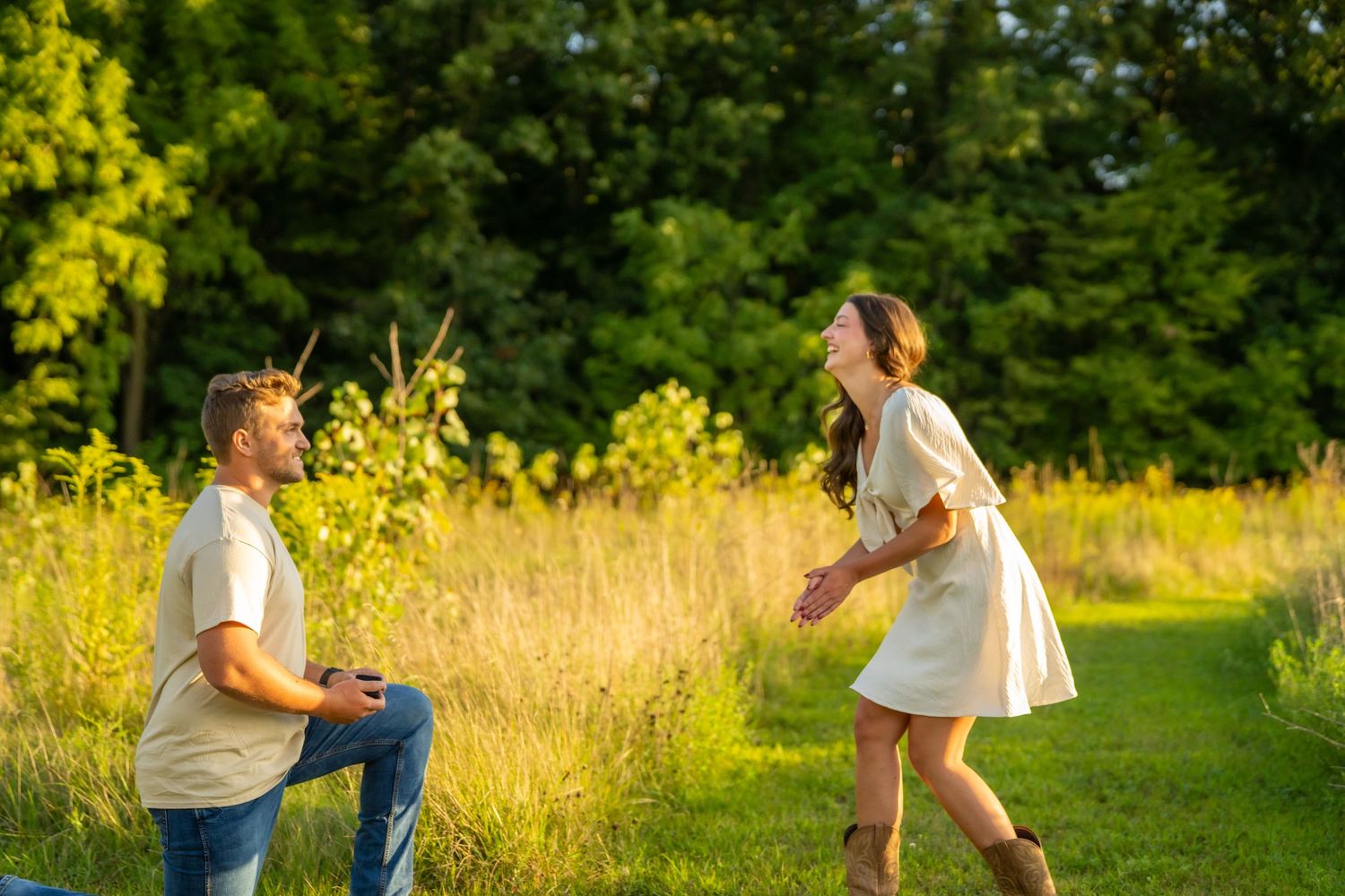 Man on one knee proposing as woman laughs with joy on a sunlit grassy meadow path.