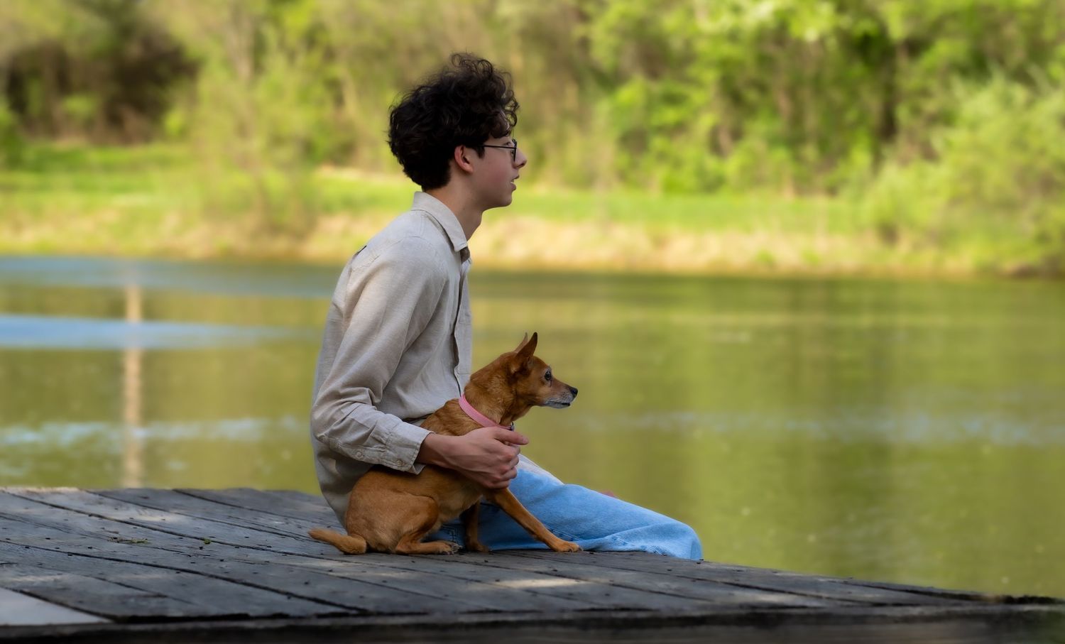Young man sitting on a wooden dock holding a small brown dog by a lake.