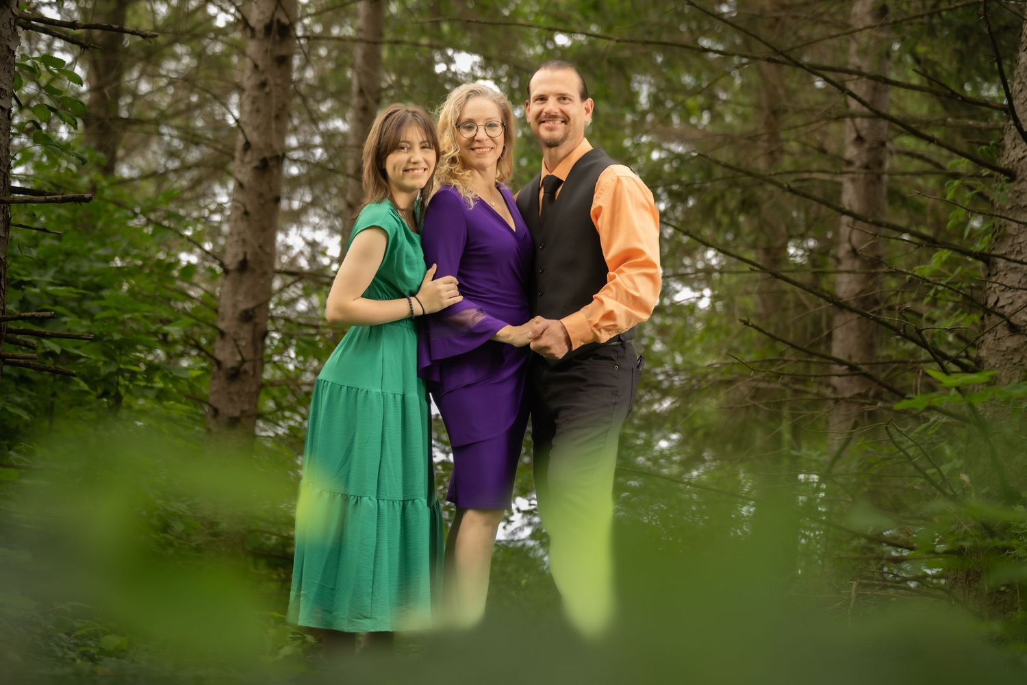 Family of three smiling together outdoors in colorful formal attire among green trees.