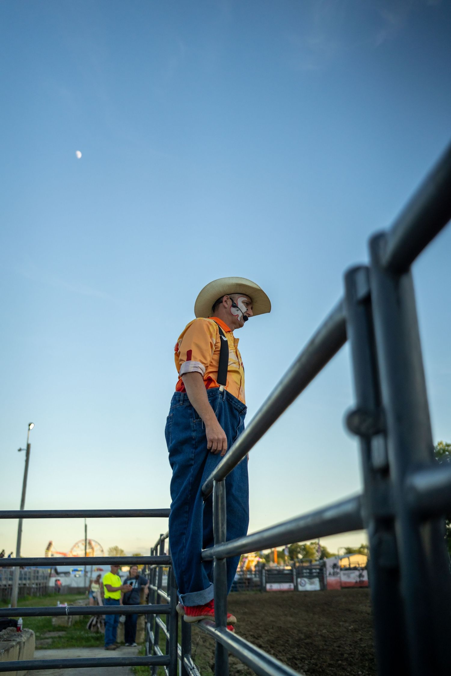 A rodeo clown in orange shirt, overalls, and cowboy hat stands on metal fence rails at dusk.