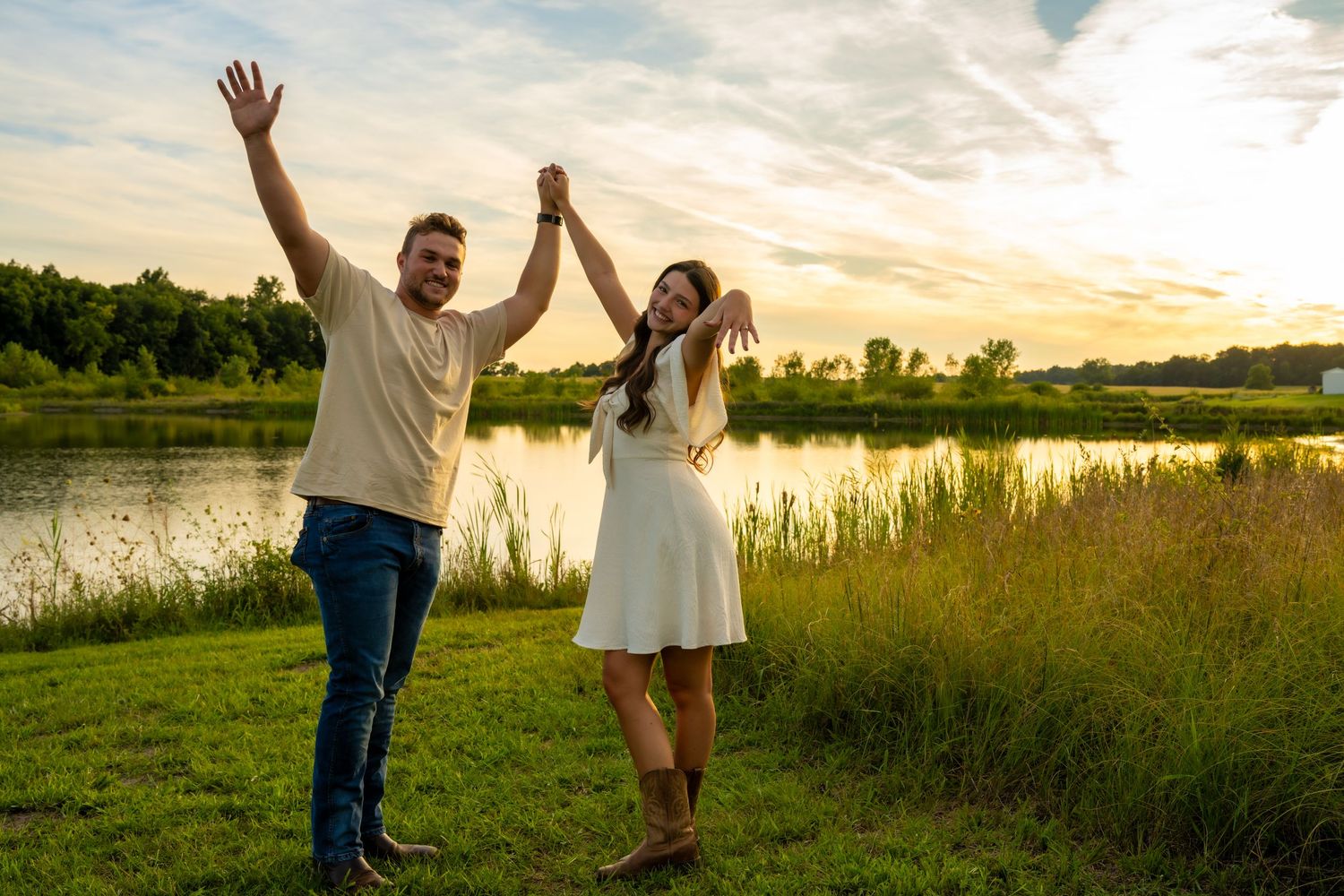 Newly engaged couple celebrating with raised hands near a scenic lake at sunset.