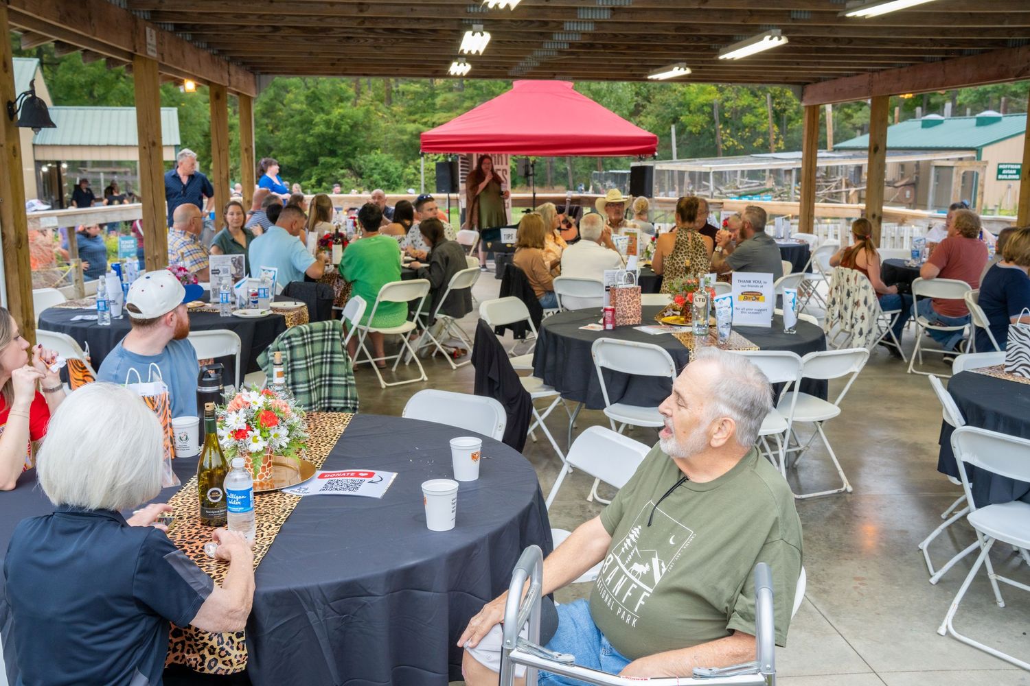 Large group of people seated at round tables under a pavilion enjoying drinks at an outdoor community gathering.