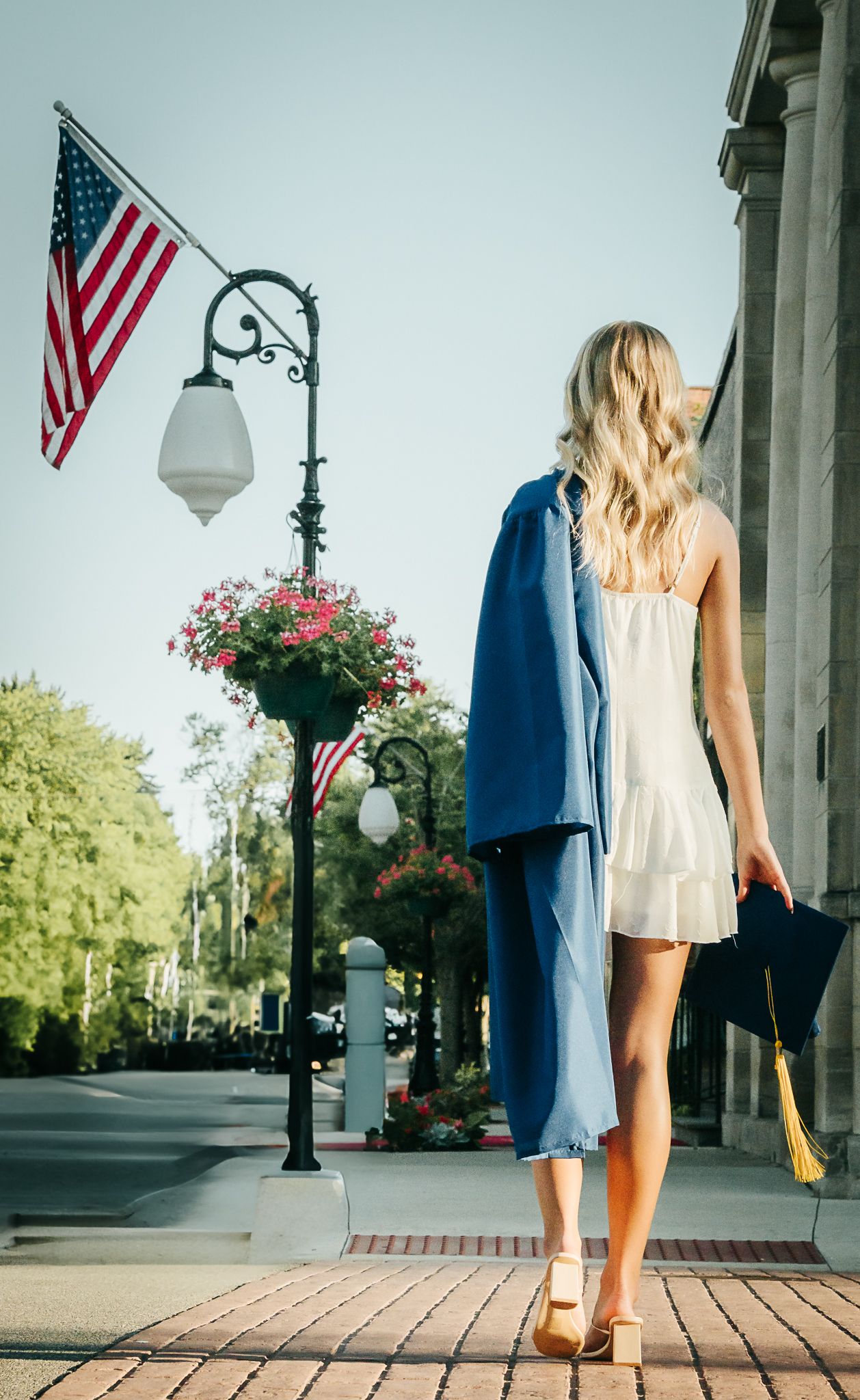Blonde woman in white dress carrying blue graduation cap and gown near American flag.