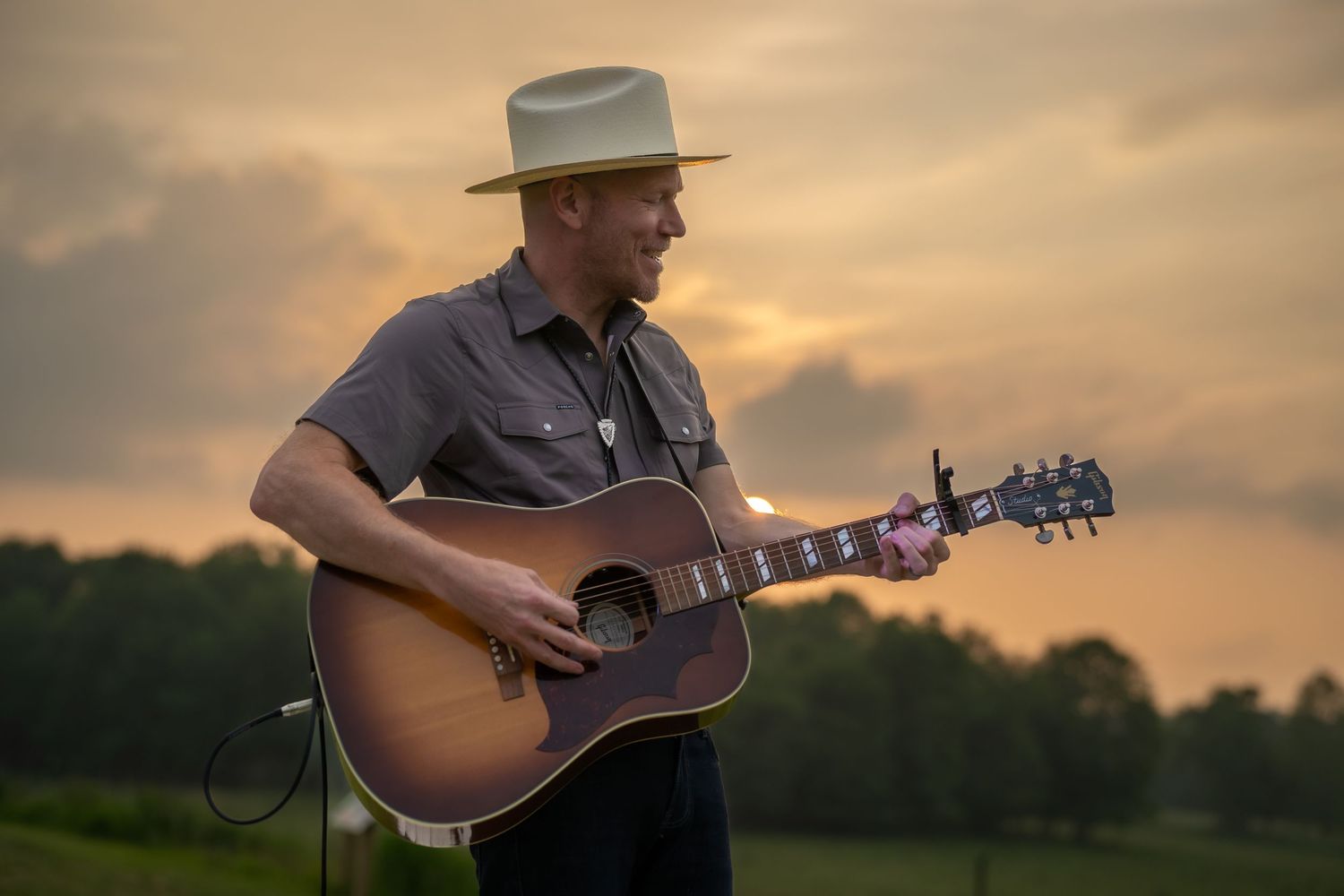 Male musician in a white hat playing acoustic guitar outdoors during a golden sunset over green farmland.