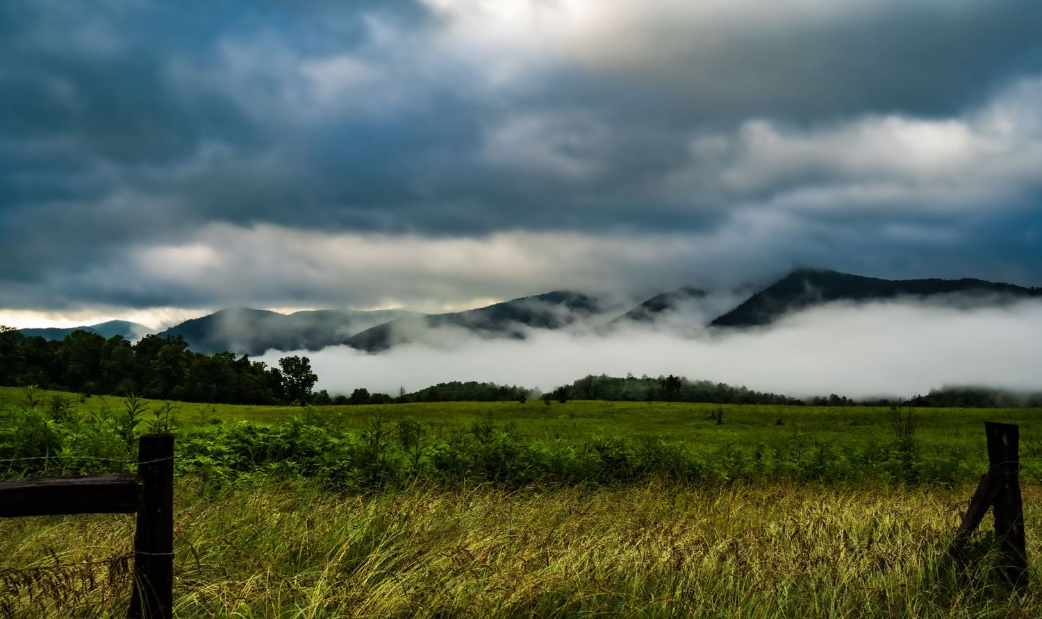 Low-lying fog blankets mountain ridges beyond a green meadow with wooden fence under stormy sky.